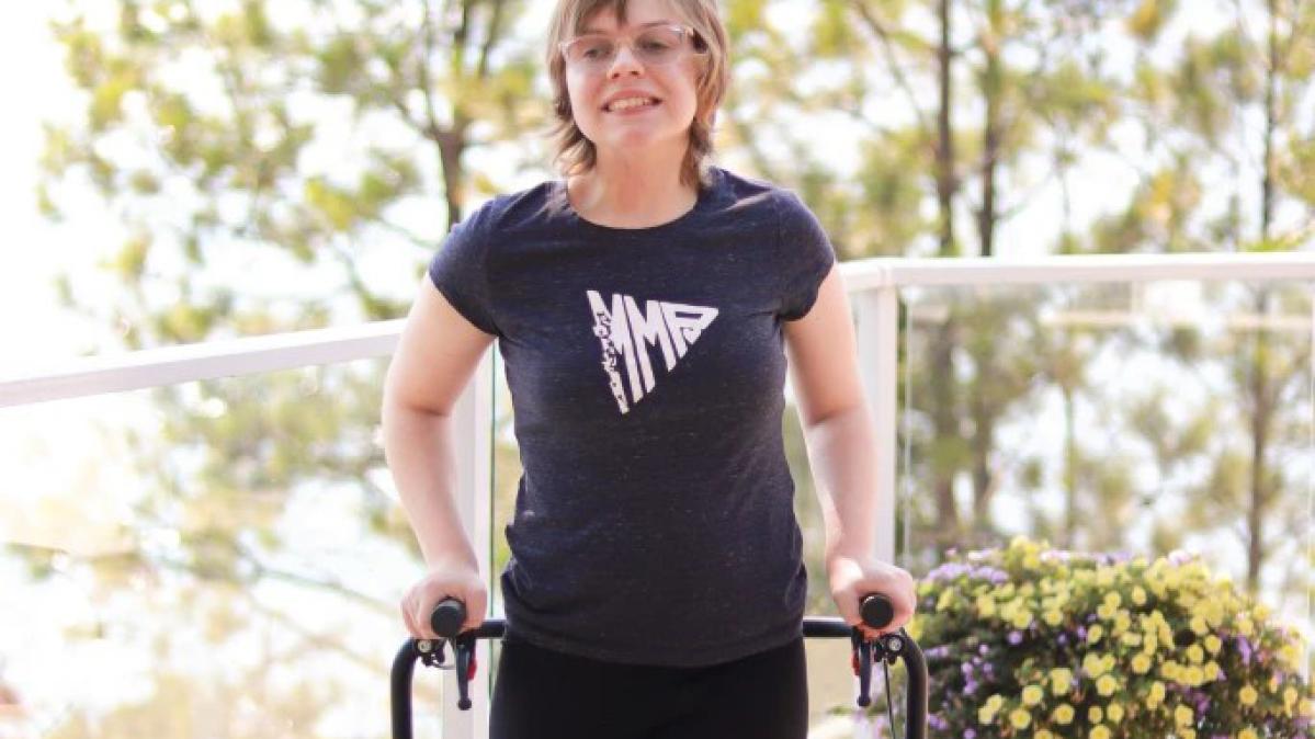 A young woman Caucasian women with short blond hair and glasses stands on a deck with glass panels and white rails behind her. She wears a black t-shirt and black pants, holds on to a blue mobility walker and smiles broadly. 