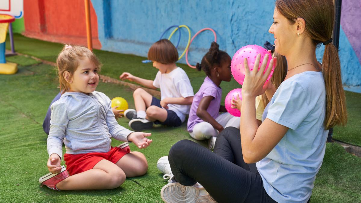 Preschool children sit on the floor and play with a ball with a young childcare worker.