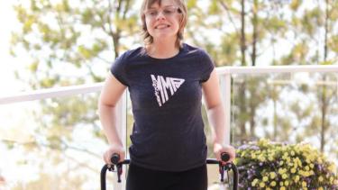 A young woman Caucasian women with short blond hair and glasses stands on a deck with glass panels and white rails behind her. She wears a black t-shirt and black pants, holds on to a blue mobility walker and smiles broadly. 