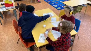 Four elementary school students sit on orange chairs around a yellow table in a colourful, warm classroom working on writing. 