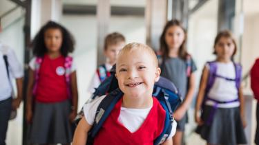 Smiling blond boy walking in a school hallway followed by other smiling young students.