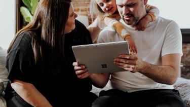 A woman, man and child look at a laptop inside a bricked-walled apartment