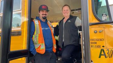 A bus driver wearing an orange reflective vest and a bus driver wearing a navy vest stand inside the doorway of a school bus on the steps.