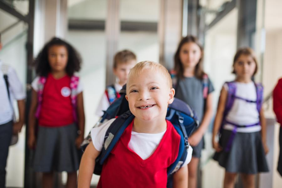 Smiling blond boy walking in a school hallway followed by other smiling young students.