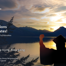Silhouette of a person in cap and gown with arms raised holding a diploma, in front of a lake and mountains at sunset